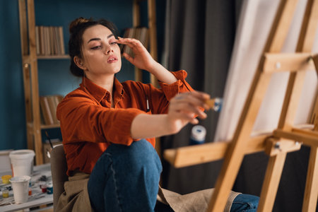a concentrated Brazilian woman artist with paint on her hands works on drawing a picture on canvas sitting in front of her, applying a brushstroke on a stool. home studio. hobby and vacation conceptの写真素材