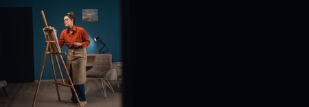 attractive Latina woman in glasses and an apron holds palette and poses with brush, artist stands near easel in the workshop. Art studio, creativity, hobby, activity for the soul. Banner. copy space.の写真素材