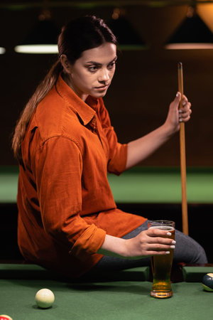 Portrait of young beautiful woman drinking beer sitting on billiard table