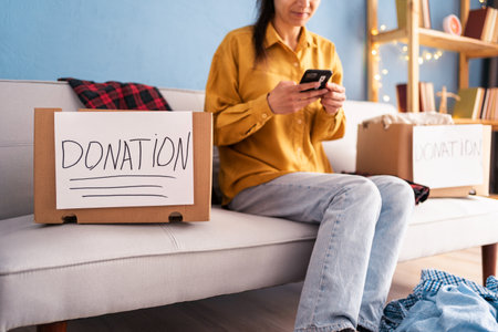 Woman packing donation boxes at home uses smartphone contacts charity centerの写真素材