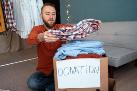Man sit on floor near big cardboard box full of old clothes, stuff used apparels is packing for donation. Humanitarian aid, charity and benevolence conceptの写真素材