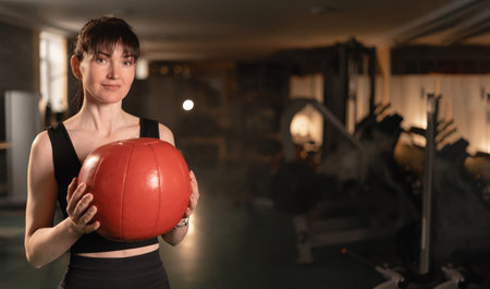 Portrait of mid adult woman looking at camera while holding heavy medicine ball over gym background. Strong woman with heavy ball after cross training workout.の写真素材