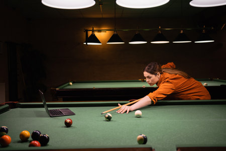 Beautiful girl learning to play billiards watching online video lesson of playing billiards using laptop on pool tableの写真素材