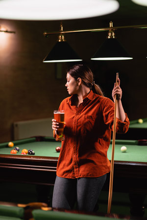 Playing billiards in a bar. Attractive girl with a cue in her hands drinks a glass of beer and...