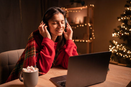 Portrait of rejoicing woman listening to christmas music rejoicing with new year atmosphere wearing santa hat sitting at table at home indoorsの写真素材