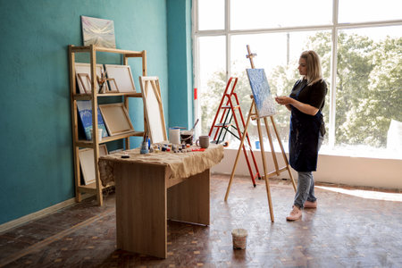Young female artist in her modern creative studio with canvases and a large window, creating a painting with smears of multicolored paint using a palette knife. Hobby, work, art and life conceptの写真素材
