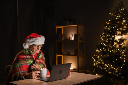 Santa Claus is watching a movie online using a laptop, sitting at the table wrapped in a checkered blanket and drinking cocoa with marshmallows. Holiday concept for christmasの写真素材