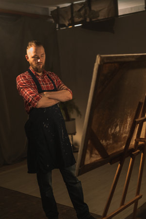 Thoughtful professional male artist wearing apron, arms crossed while looks at the canvas. Authentic creative studio with large canvasの写真素材