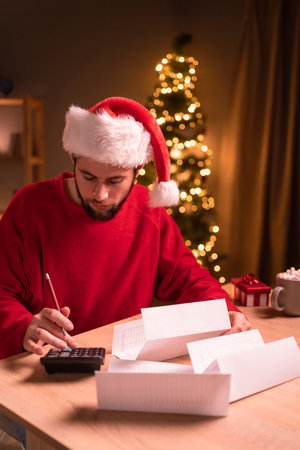 Man wearing red sweater and hat using calculator and looking at bill with the christmas tree in the backgroundの写真素材
