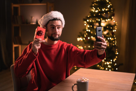 Young happy man having video call over smartphone and showing gift box at camera while celebrating Christmas alone at home.の写真素材