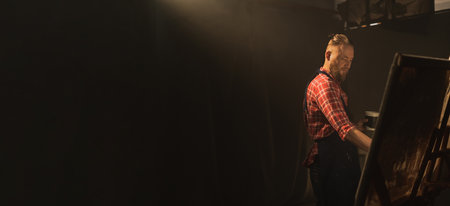 Talented male artist creates modern masterpiece of the painting using paint brush. Working in artist workshop. Dark creative studio with large canvas. Side viewの写真素材