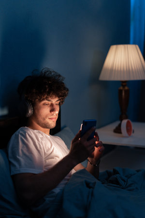 Handsome mobile addict man using smartphone browsing the internet for watching movie or virtual meeting video call online with girlfriend on the bed, sleep time at night.の写真素材