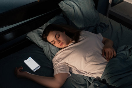 Mobile use addiction concept. Addicted to social media young woman falling asleep with smart mobile phone at night in bed with screen light on. Girl sleeping in dark bedroomの写真素材