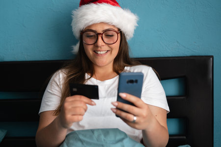 Online payment, Woman holding a credit card and using smart phone for online christmas shopping sitting resting in bedroom. Happy girl with glasses buying holiday gifts onlineの写真素材