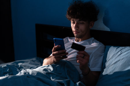Handsome man sitting on the bed and using smart phone to pay with credit card at night at home. Credit bank card for online shop, order delivery, booking tour, relax at home in bedroomの写真素材