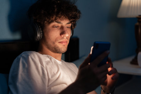 Portrait of a serious guy listening to music with headphones and smart phone lying on the bed in the night at home, watching movie onlineの写真素材