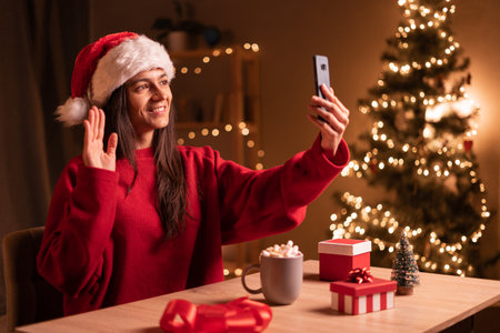 Woman waving during a smart phone video call in christmas sitting at desk at homeの写真素材
