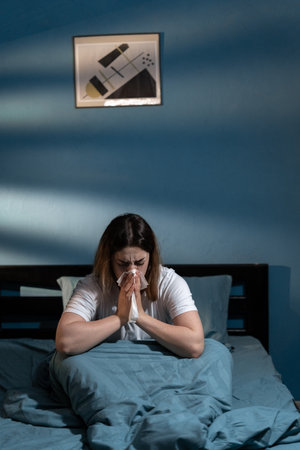 Sick girl wake up from sleep at night sneezing on bed. Attractive young woman suffer from allergy, put tissue cover her nose while sneezes during bedtime in house.の写真素材