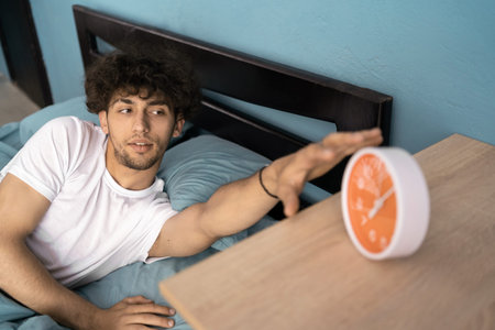 Happy young man 20s lies in bed under blanket duvet on pillow turn off clock alarm rest relax indoors at home. Good mood morning bedtime concept.の写真素材