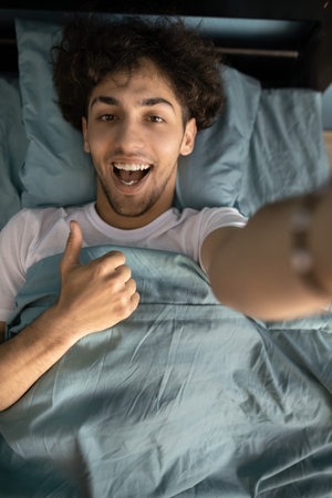 Male person lying in bed making photos with thumb up in room indoorsの写真素材