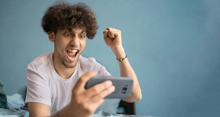 Portrait of a happy young man in white t-shirt looking at smartphone and celebrating win victory at home interiorの写真素材