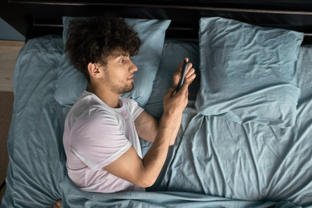 Internet addiction and mobile abuse. Man in bed chatting uses smartphone early morning. World in phone.の写真素材