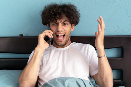 Young arab sitting in bed talking on cellphone and shouting loudly into phone, nervous stressの写真素材