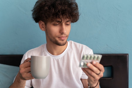 Attractive young Arab men hold pills and cup of water in bedroom interior. Disease treatment, health care, medicine, vitamins and therapyの写真素材