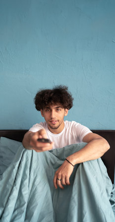 Young handsome man watches TV while sitting in bed in the morning, uses the remote control