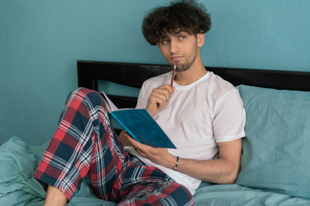 Cropped shot of arab man sits wears pajamas, makes notes in diary, focused aside, domestic interior. Male blogger creates publication for blog in notepadの写真素材