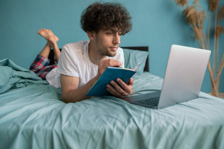 Young arab guy student studying virtual course, learning distance class taking web training, looking at laptop computer, writing notes sitting on bed at home.の写真素材