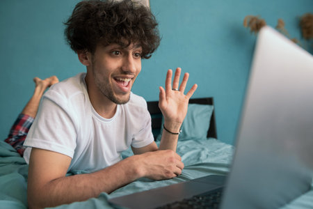 Happy smiling young man has video call using laptop computer in bed at home bedroom. Technology,...