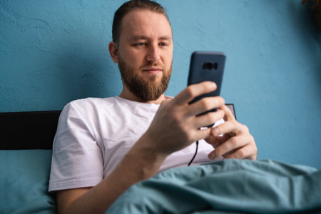 Portrait of smiling young freelance businessman using mobile phone checking social media network sitting on bed at home. Online digital communicationの写真素材