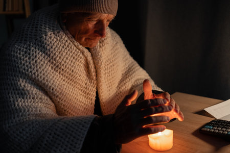 Freezing old man in winter clothes warms his hands over candle as energy blackouts cause electricity outages.の写真素材