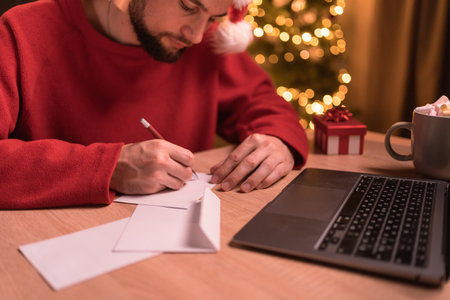 Bearded man in Santa hat writing greeting cards at home office during christmasの写真素材