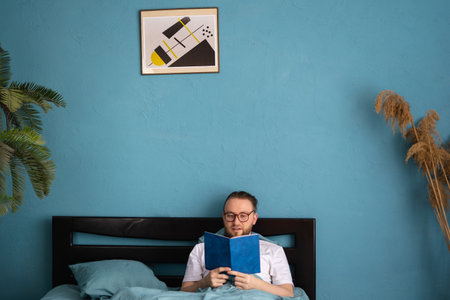 Happy man wear t-shirt lying in bed and read book, study spend time in bedroom, wake up has good mood day.の写真素材