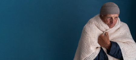 Portrait of senior man freezing at home in winter over blue background. Elderly man wearing hat and wrapped in blanket suffering from low temperature, in house with bad broken thermostatの写真素材