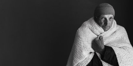 Black and white photo of unwell old man in hat and blanket over black background. Unhealthy senior struggle from chill freeze at home. No heating concept.の写真素材