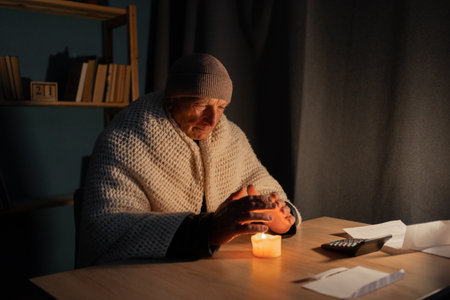 Freezing old man in winter clothes warms his hands over candle as energy blackouts cause electricity outages.の写真素材