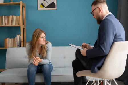 Psychologist taking notes during therapy session with sad young woman in his office.