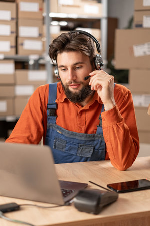 Male worker with headset working in on-site office of a warehouse using laptop computer consulting clients.の写真素材