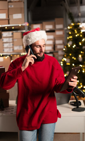 Logistics, shipping and warehouse worker on the phone with tablet. Man with smartphone in delivery, freight and international distribution business in storehouse on Christmas.の写真素材