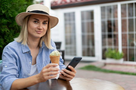 Woman eating ice cream using smartphone in street cafe. Portrait of a girl with ice cream browsing through social media or messaging outdoorsの写真素材
