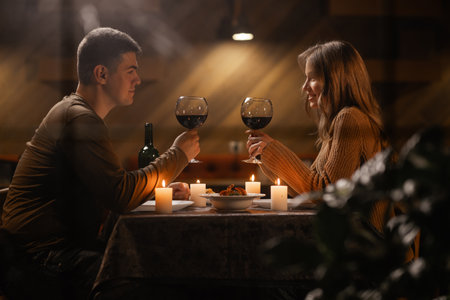 Trough window view on young couple in love drinking wine, celebrating Valentines day dining at home, having romantic dinner date with candlesの写真素材
