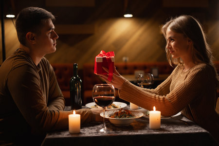 Happy young couple celebrating Valentines day having romantic dinner at home, woman giving gift box for beloved man making present surprise on candle light dateの写真素材