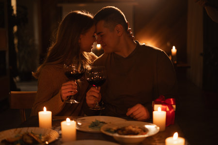 Happy young couple in love hugging holding glasses having romantic dinner date with candles in dark light, drinking wine, celebrating Valentines day at homeの写真素材