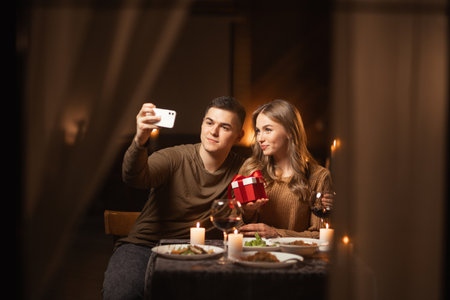 Valentines selfie. Happy young couple making self-portrait on smartphone holding gift box having date in restaurant or at homeの写真素材