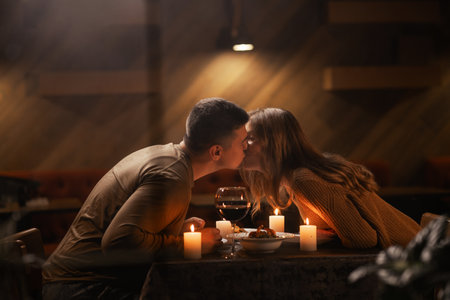 Happy young couple in love kissing, drinking red wine having romantic dinner, celebrating Valentines day, anniversary, sitting at homeの写真素材