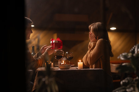 Romantic dinner for two at home, a man gives a woman a gift for a birthday or valentines day, a couple in love in the evening, sitting at a table with candles, concept of holiday giftの写真素材