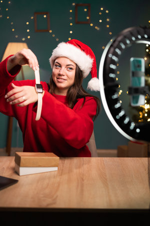 Video review. woman influencer demonstrates smart watch in front of smartphone camera, puts it on hand, writes content for her fashion blog, sitting at home in Santa hat. product advertising conceptの写真素材